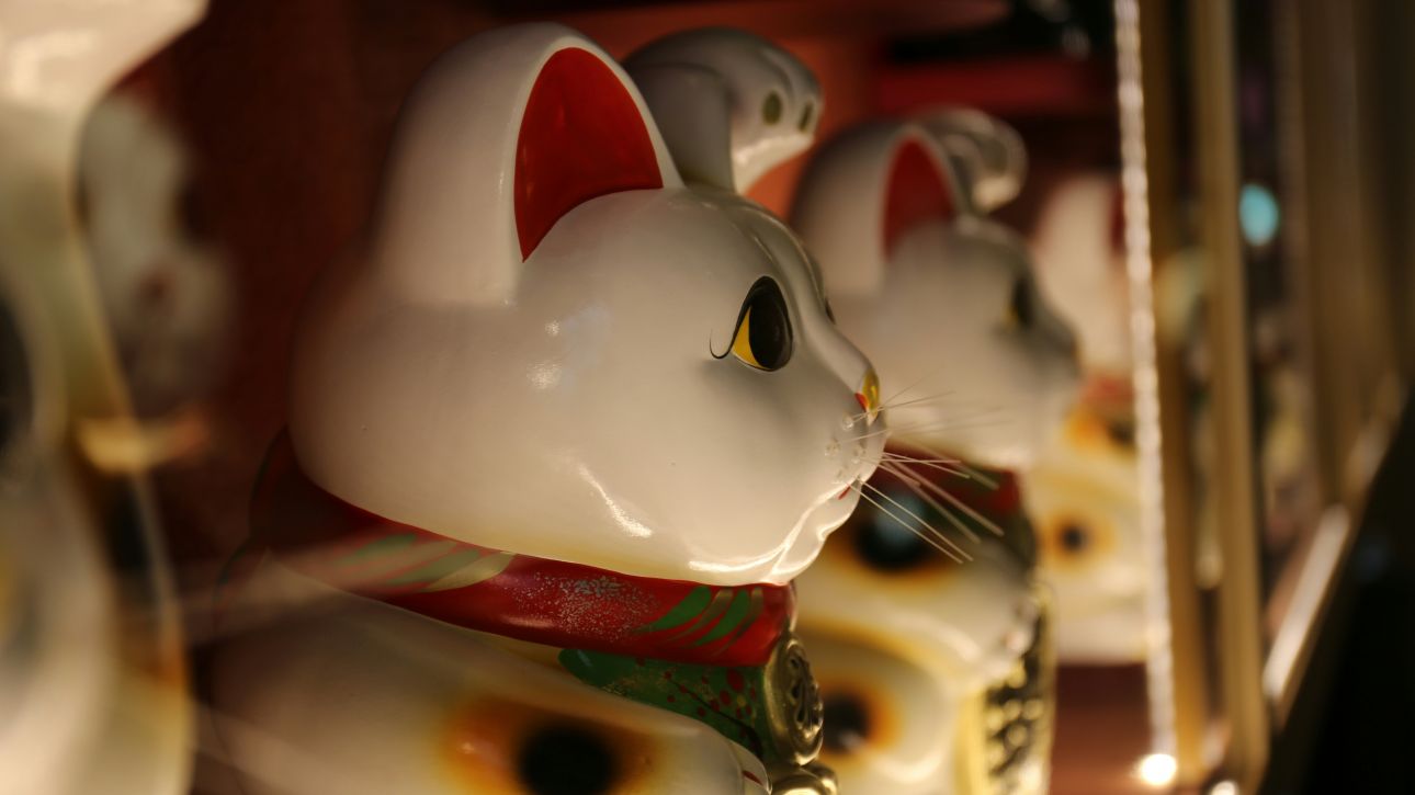 Nahaufnahme einer Maneki-neko-Statue, einer japanischen winkenden Katze, mit weiteren Figuren unscharf im Hintergrund.
